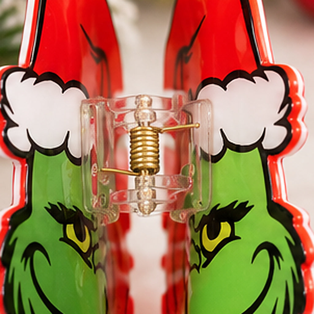A close-up of a clear plastic hair claw clip with a gold metal spring, positioned over a red and green Grinch-themed hair clipper featuring Santa hat and green face details.