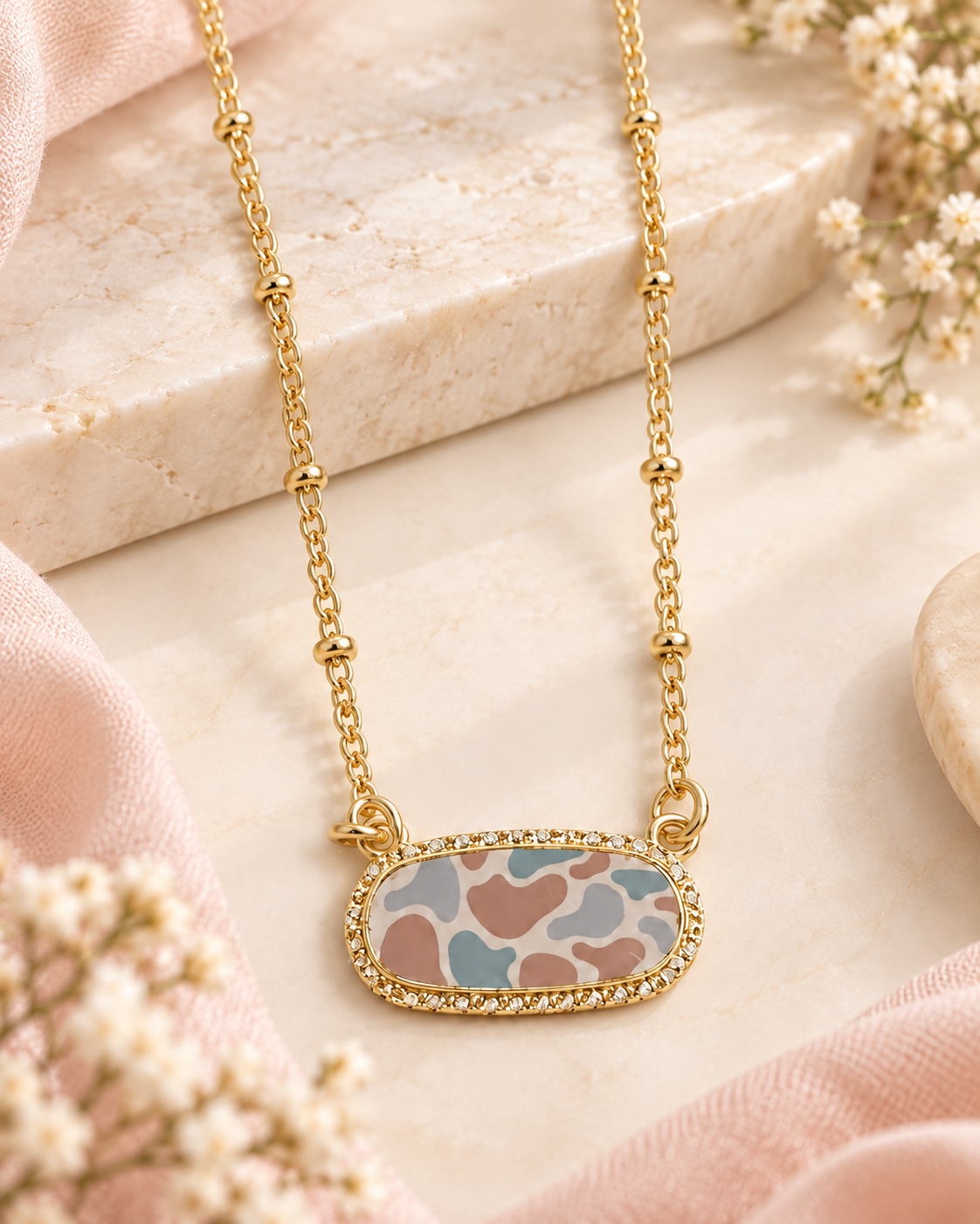 Gold chain necklace with a rectangular pendant featuring a pastel camouflage pattern in pink, blue, and beige, surrounded by a small border of clear rhinestones, displayed on a light marble slab with pink fabric and baby's breath flowers.