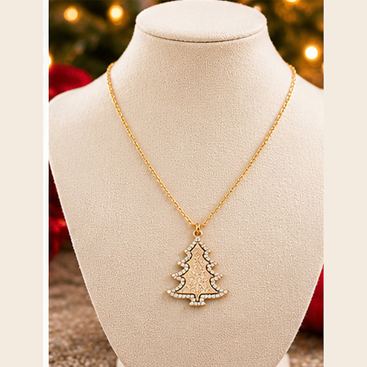 A gold necklace with a Christmas tree pendant displayed on a cream-colored jewelry bust. The pendant is outlined with small clear rhinestones and filled with a textured gold pattern.