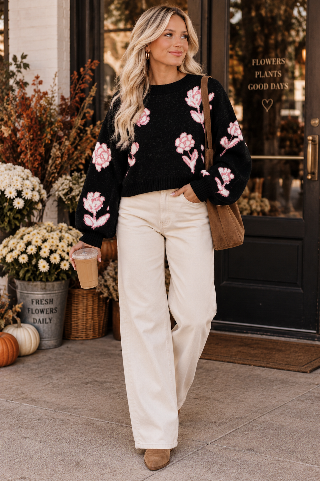A woman stands on a sidewalk in front of a storefront wearing a black knit sweater with pink floral embroidery and cream wide-leg pants. She is holding a coffee cup and wearing a brown shoulder bag.