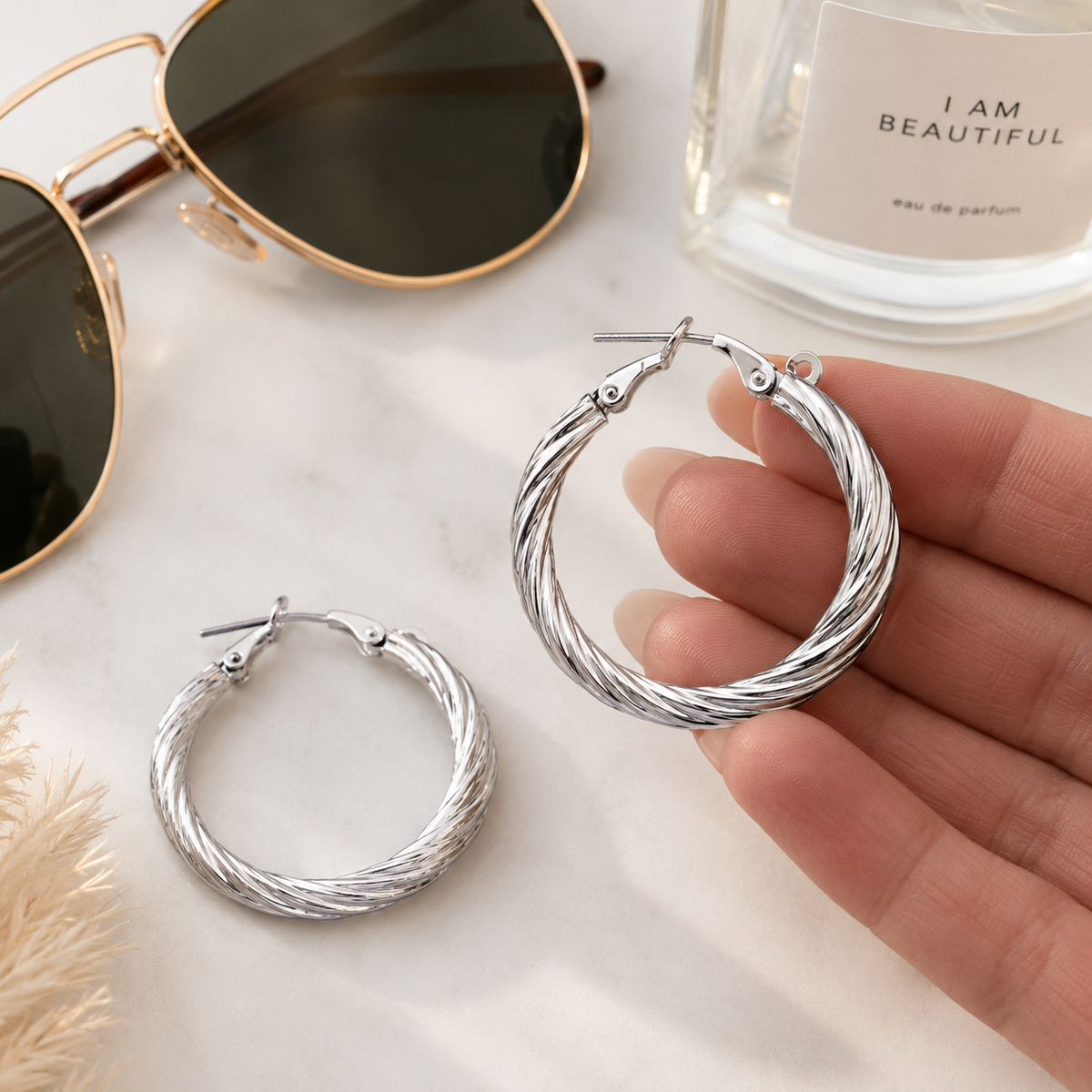 A pair of silver hoop earrings with a twisted rope texture, shown on a light marble surface with sunglasses, a perfume bottle, and a hand holding one earring.