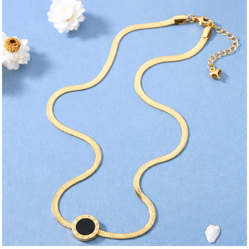 A gold snake chain necklace with a circular black and gold pendant, displayed on a textured light blue background with small white flower props.