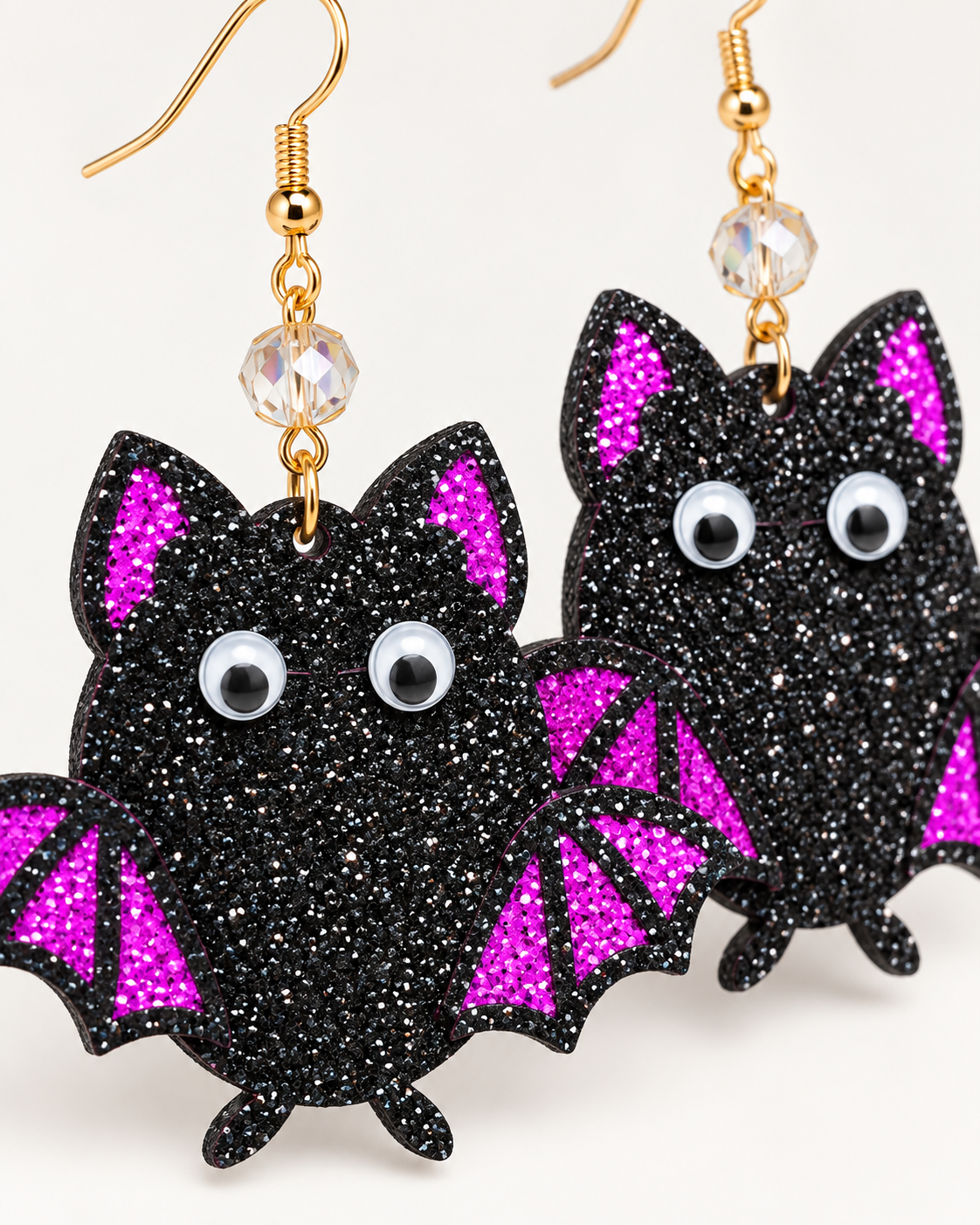 Black glitter bat-shaped dangle earrings with bright pink glitter wing and ear accents, large googly eyes, and gold fishhook wires on a plain light background.