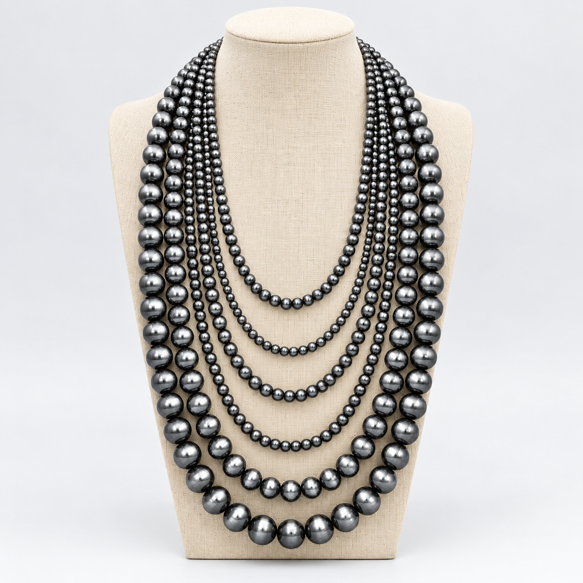 A multi-strand necklace made of large dark gray to black pearls, displayed on a beige fabric jewelry bust against a light gray background.