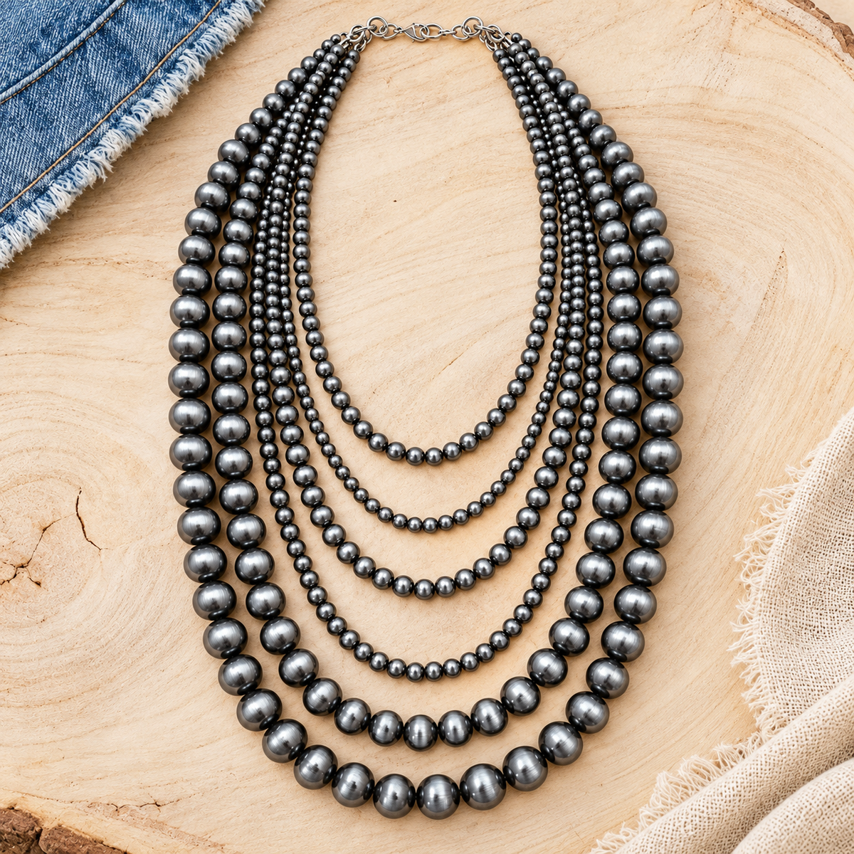 A multi-strand pearl necklace with dark gray to black pearls, styled on a light wood slice background with denim and fabric props.