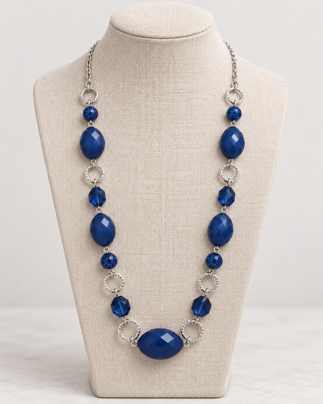 A statement necklace with deep blue faceted beads and silver-tone circular links, displayed on a beige fabric jewelry bust against a light gray background.