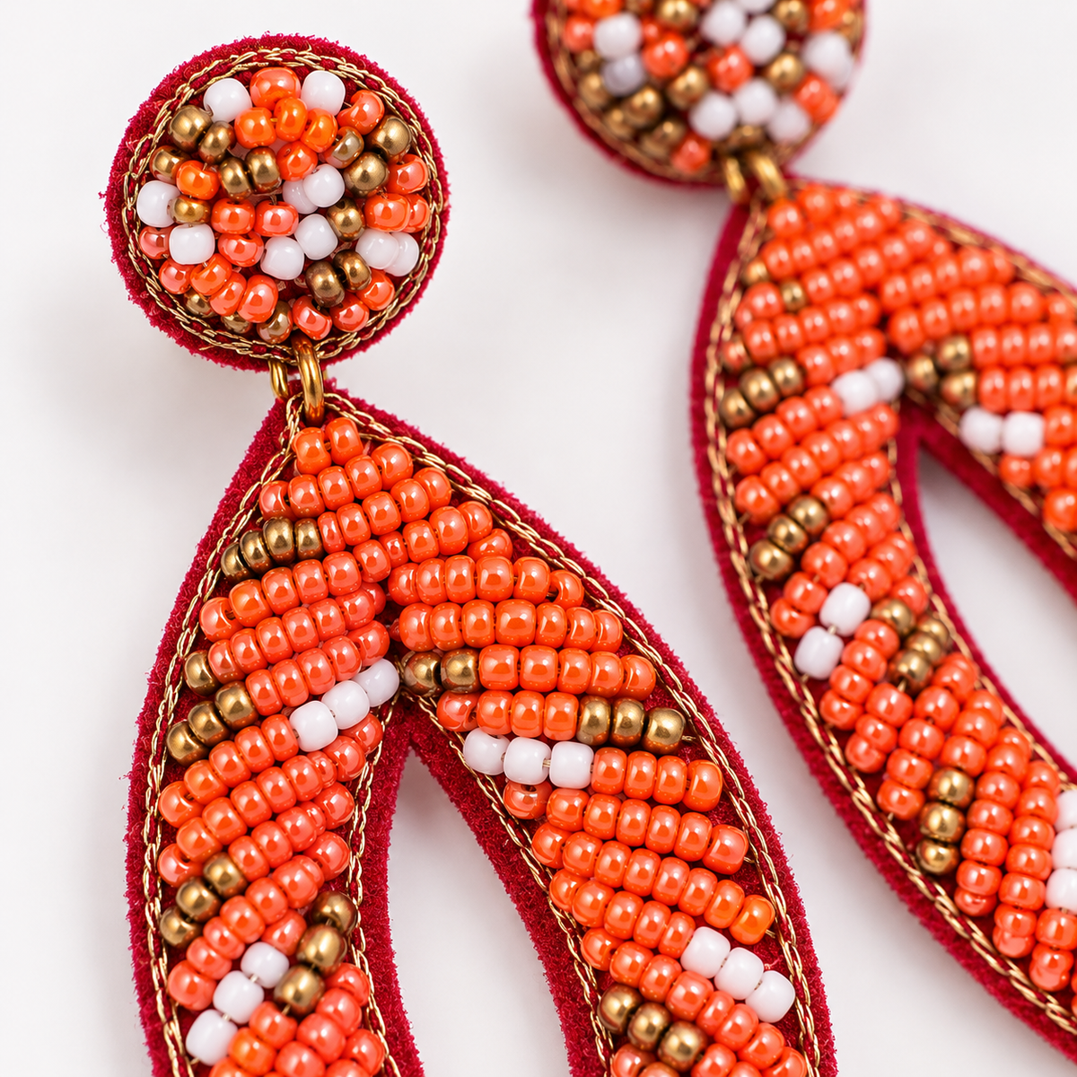 Close-up of a pair of beaded dangle earrings on a white background. The earrings feature a circular top section with orange, white, and metallic gold beads, hanging from a red fabric loop that forms a curved leaf-like drop.
