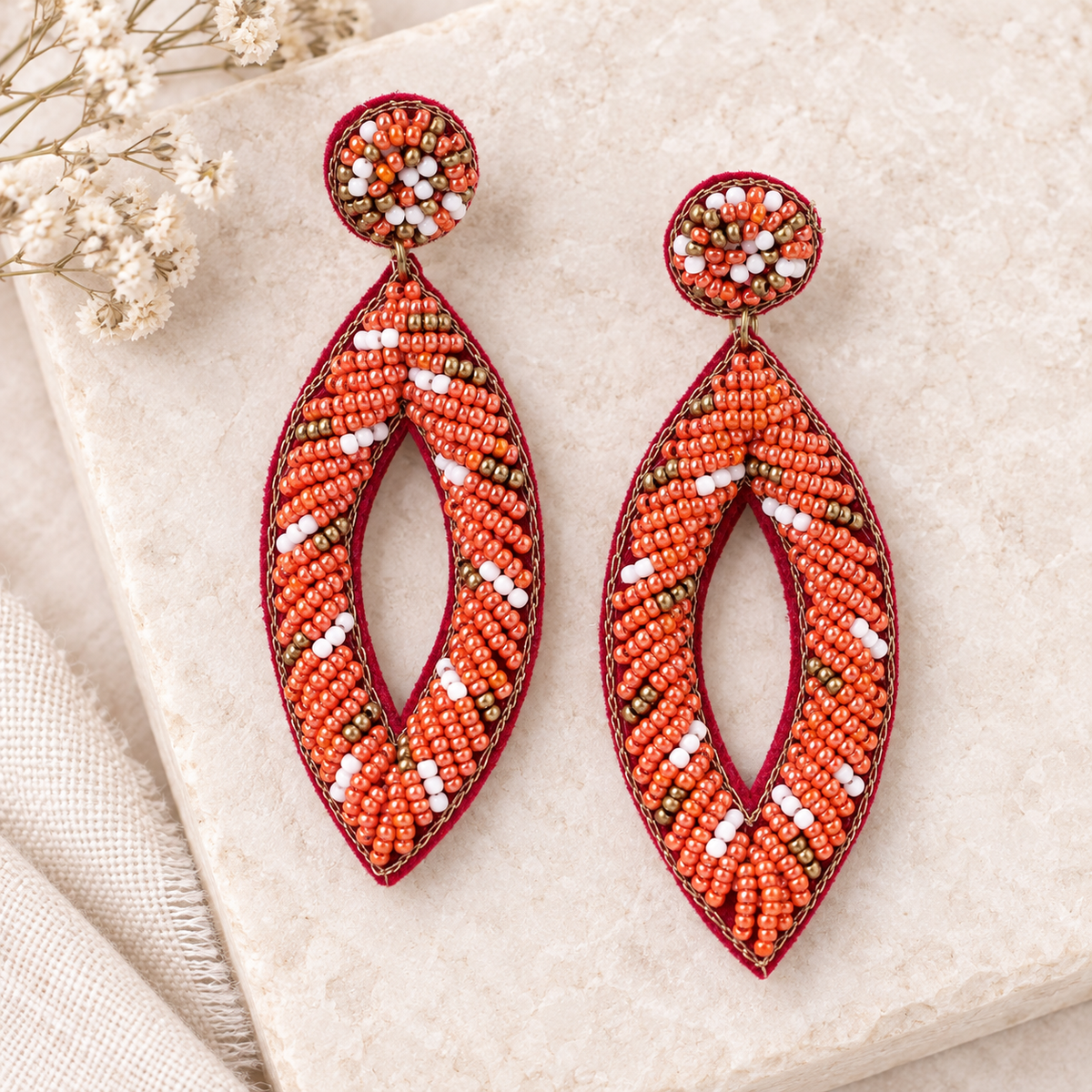 A pair of large beaded dangle earrings with a deep red border, orange and white seed beads, and a small circular bead top, displayed on a textured beige stone surface with dried flowers.