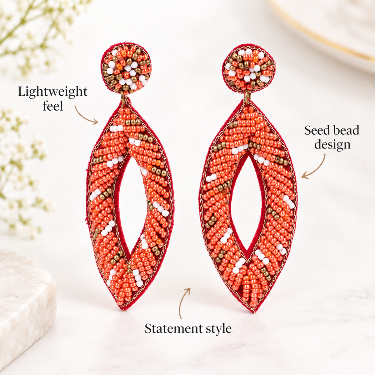 A pair of large statement earrings made from red seed beads and wrapped in red thread, displayed on a light marble surface with decorative flowers in the background. Text overlays describe the features: 'Lightweight feel', 'Seed bead design', and 'Statement style'.