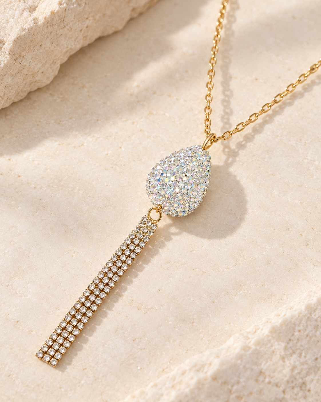A gold-tone tassel necklace with a teardrop-shaped pavé pendant, displayed on a textured beige stone surface.