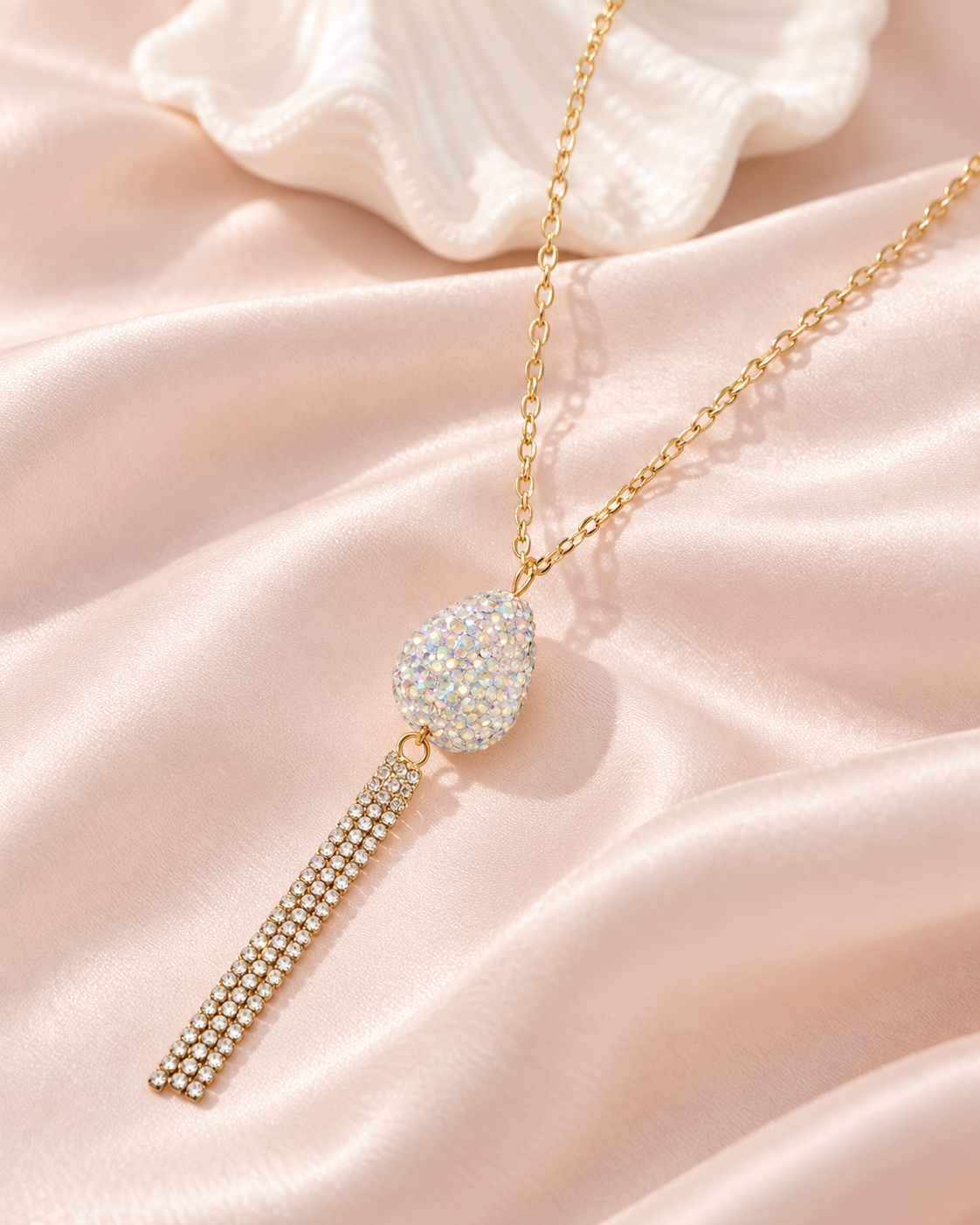 A gold chain necklace with a teardrop-shaped pavé crystal pendant and a dangling crystal-encrusted tassel, displayed on soft pink satin fabric.
