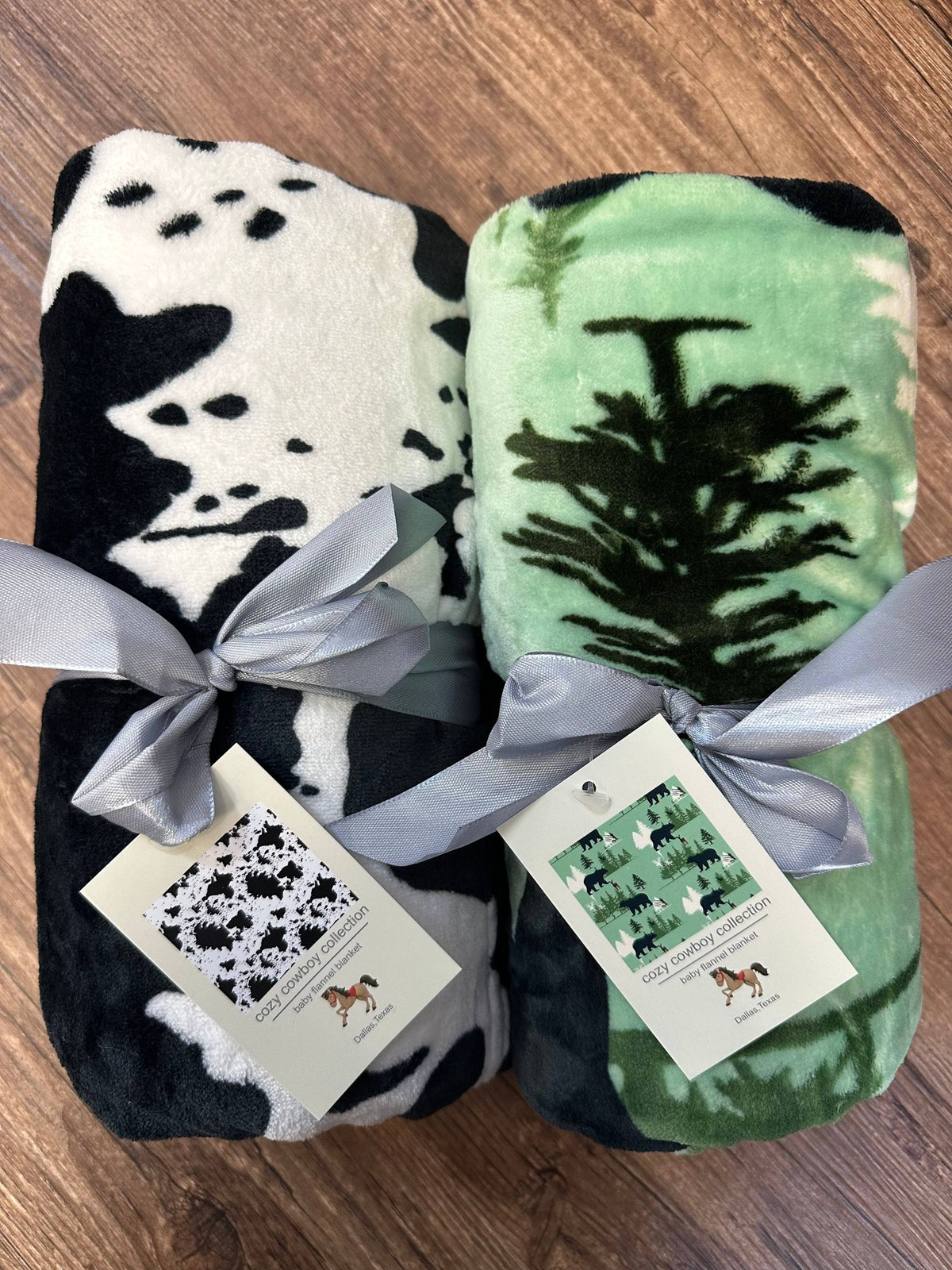 Two fleece blankets with cow print and green forest design, tied with ribbons on a wooden surface.