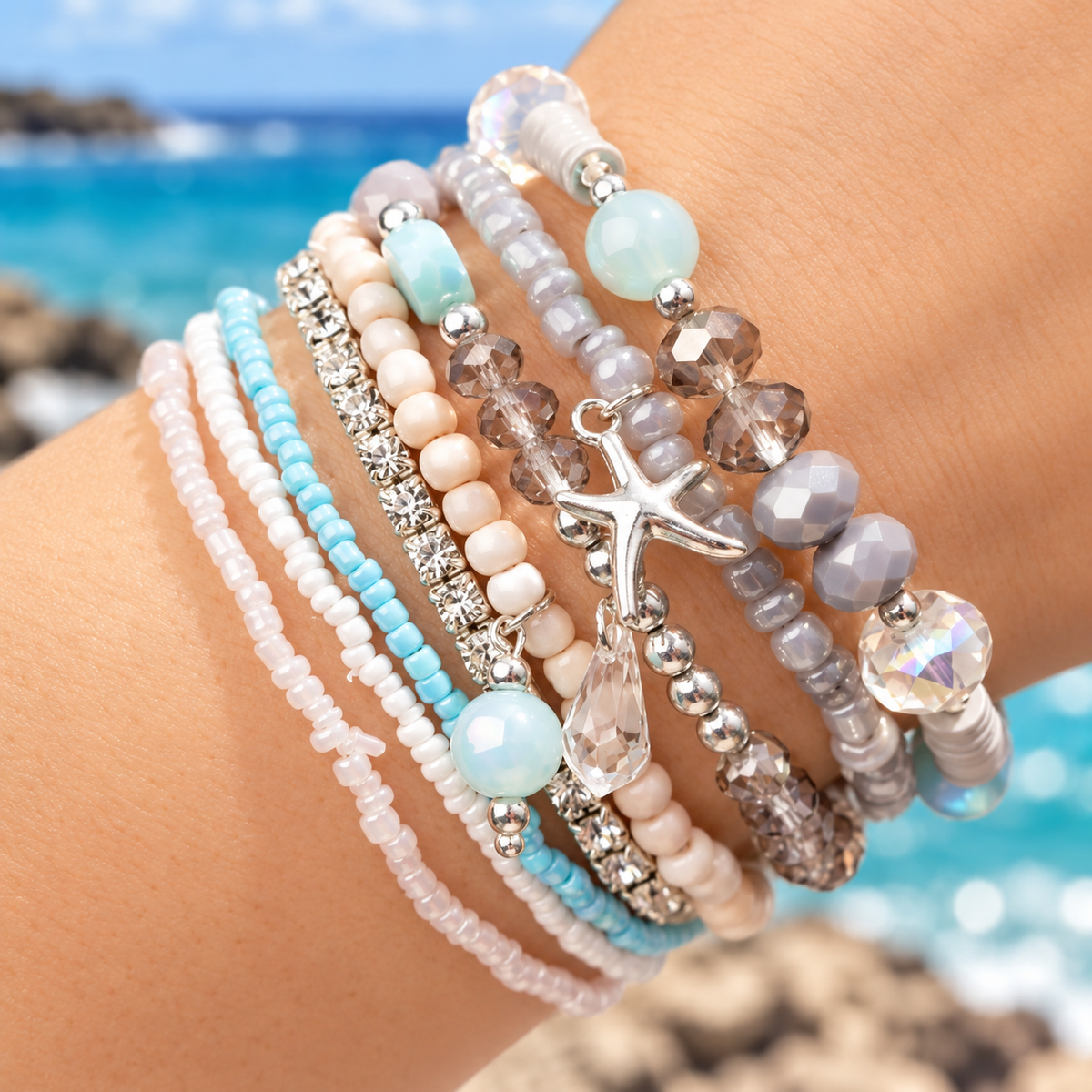 A close-up of a person's wrist wearing a stack of beaded bracelets with pastel blue, pink, white, and gray beads, faceted crystals, a silver starfish charm, and clear rhinestone accents, with a blurred beach background.