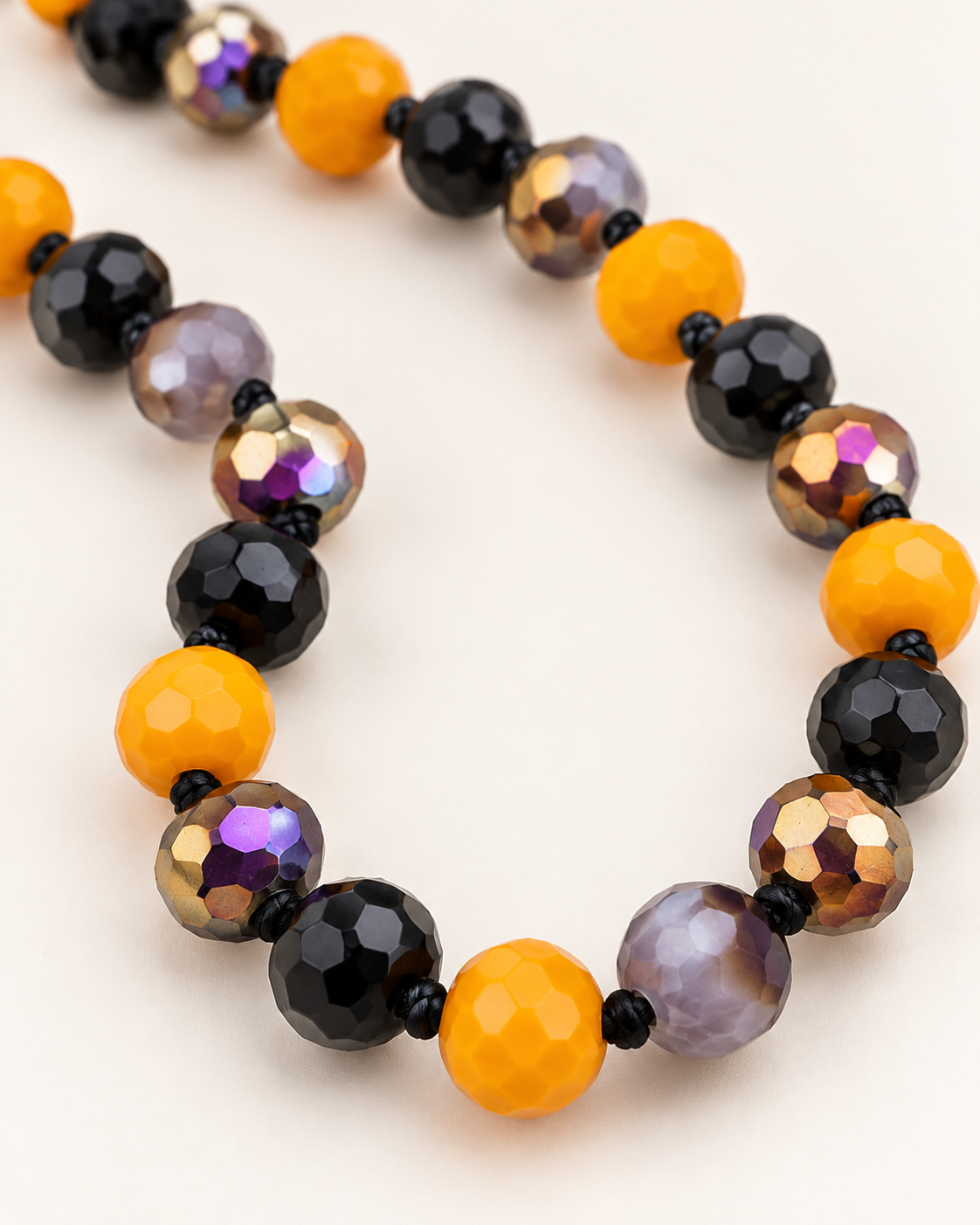 A beaded necklace with alternating matte orange, faceted black, and faceted iridescent purple beads on a light cream background.