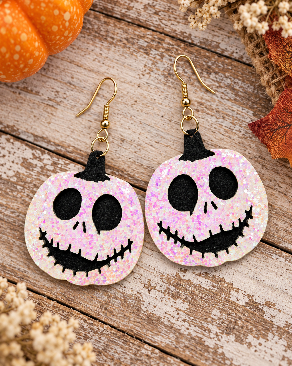 A pair of Halloween jack-o'-lantern dangle earrings with a pink glittery pumpkin face and gold-tone hooks, displayed on a rustic wooden surface with autumn decorations.