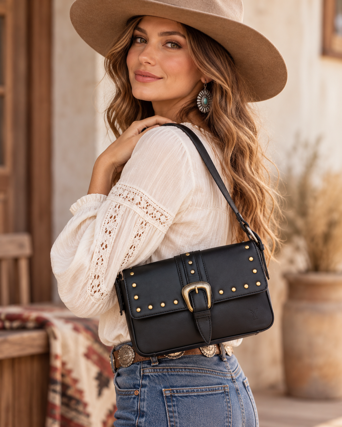 A woman wearing a wide-brim tan hat, cream boho blouse, and blue jeans carries a black faux-leather shoulder bag with gold studs and a gold buckle. The background is a warm, rustic, desert-style interior.