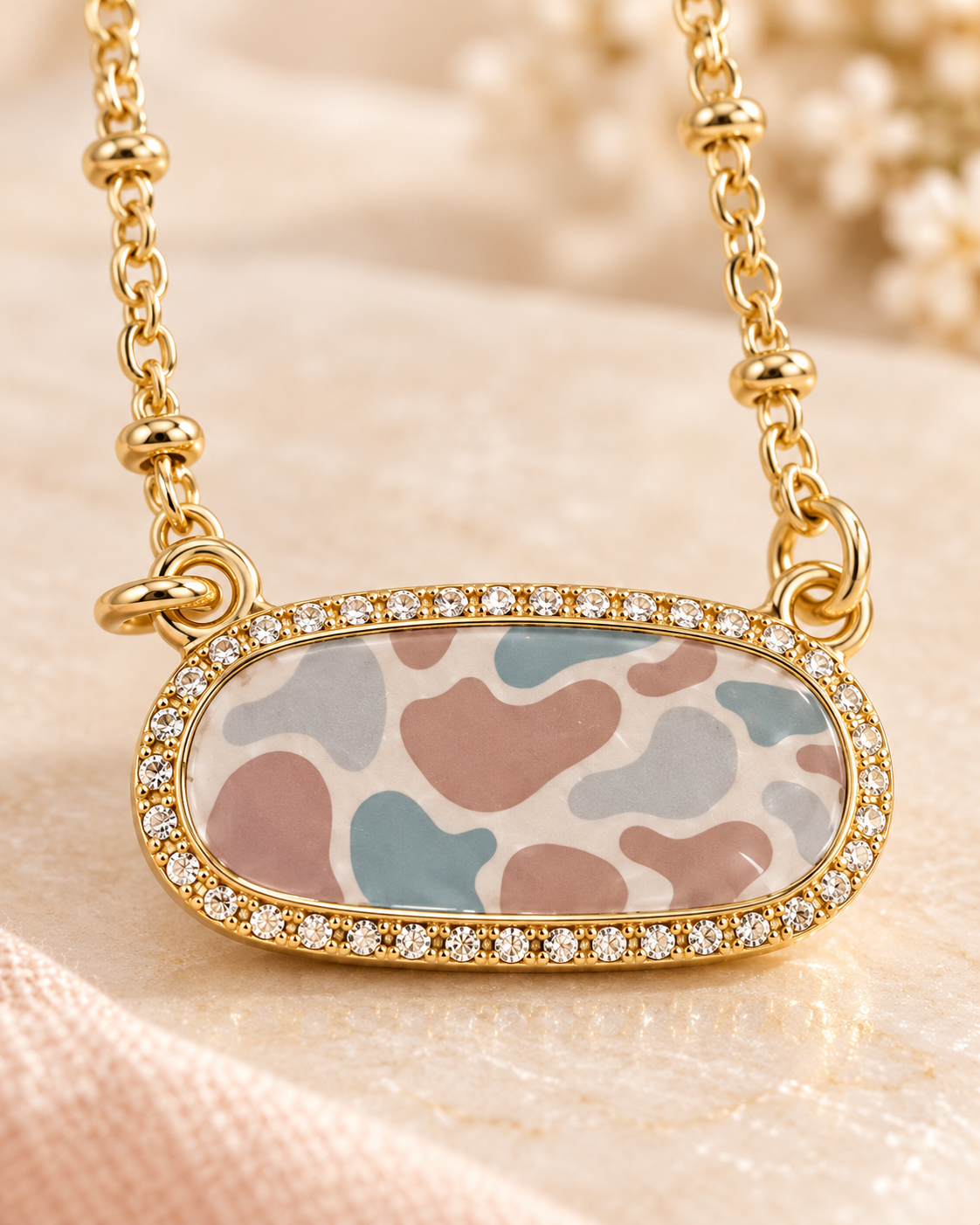 Gold-tone oval pendant necklace featuring a pastel camouflage design in blush pink, dusty blue, and gray, framed by sparkling clear rhinestones and suspended from a delicate gold-tone beaded chain.