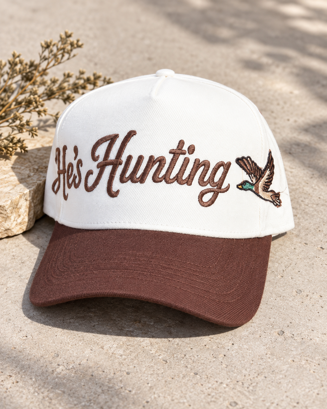 A white and brown baseball cap with 'He's Hunting' embroidered in brown script on the front, next to an embroidered mallard duck. The cap is styled outdoors on a concrete surface with a small stone and dried leaves.