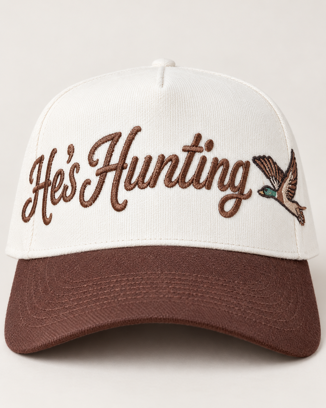 A baseball cap with an off-white crown and a brown visor, featuring the embroidered text 'He's Hunting' in brown script and a small brown bird with a green chest on the side.