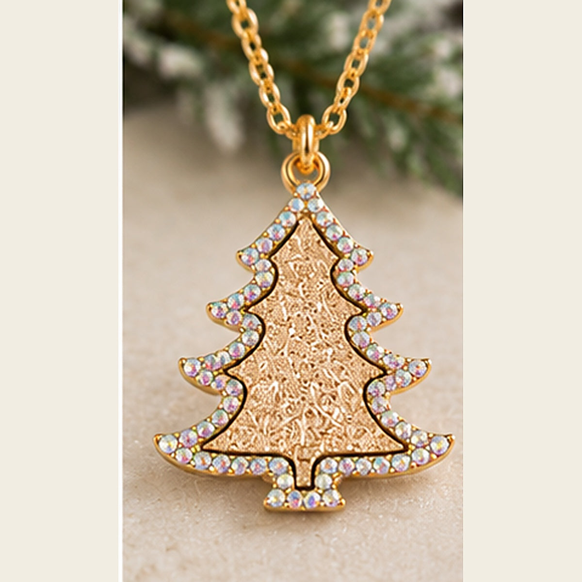 A gold Christmas tree pendant necklace adorned with iridescent rhinestones along the edges, shown on a beige surface with a blurred green background.