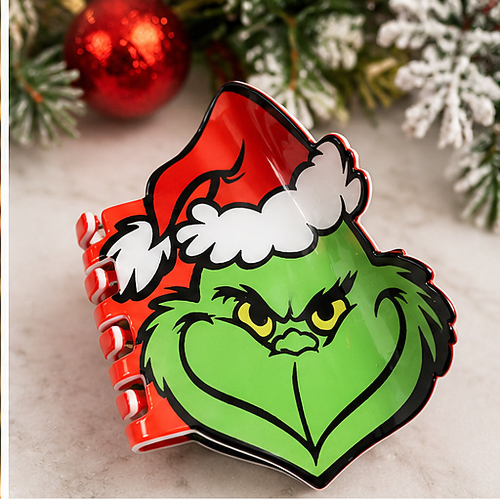 A spiral-bound notebook featuring a glossy graphic of the Grinch wearing a red Santa hat with white trim. The notebook is styled on a light marble surface with festive holiday decorations in the background, including pine branches, a red ornament, and white snow-dusted foliage.