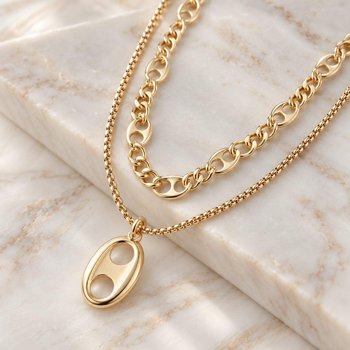 A gold-toned double-strand necklace featuring a short chain with a polished oval pendant and a longer curb chain draped above it, displayed on a cream-colored marble surface.