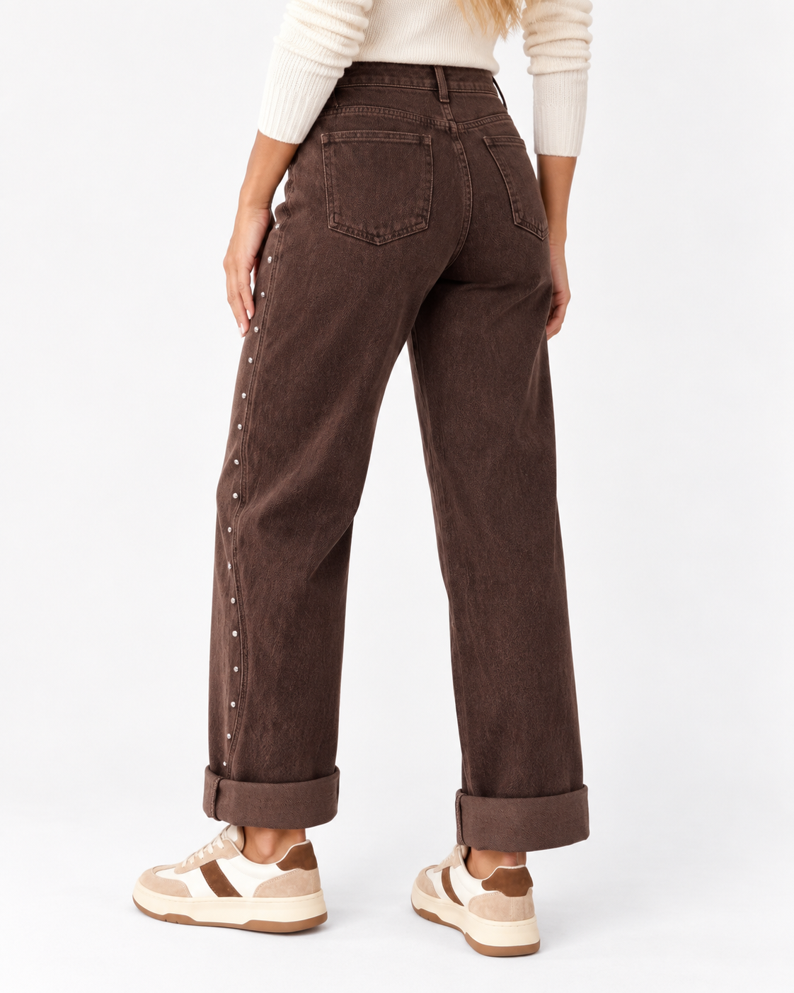 A model shown from the back wearing dark brown wide-leg jeans with silver-tone studs down the side, paired with a cream ribbed sweater and beige sneakers, against a plain white studio background.