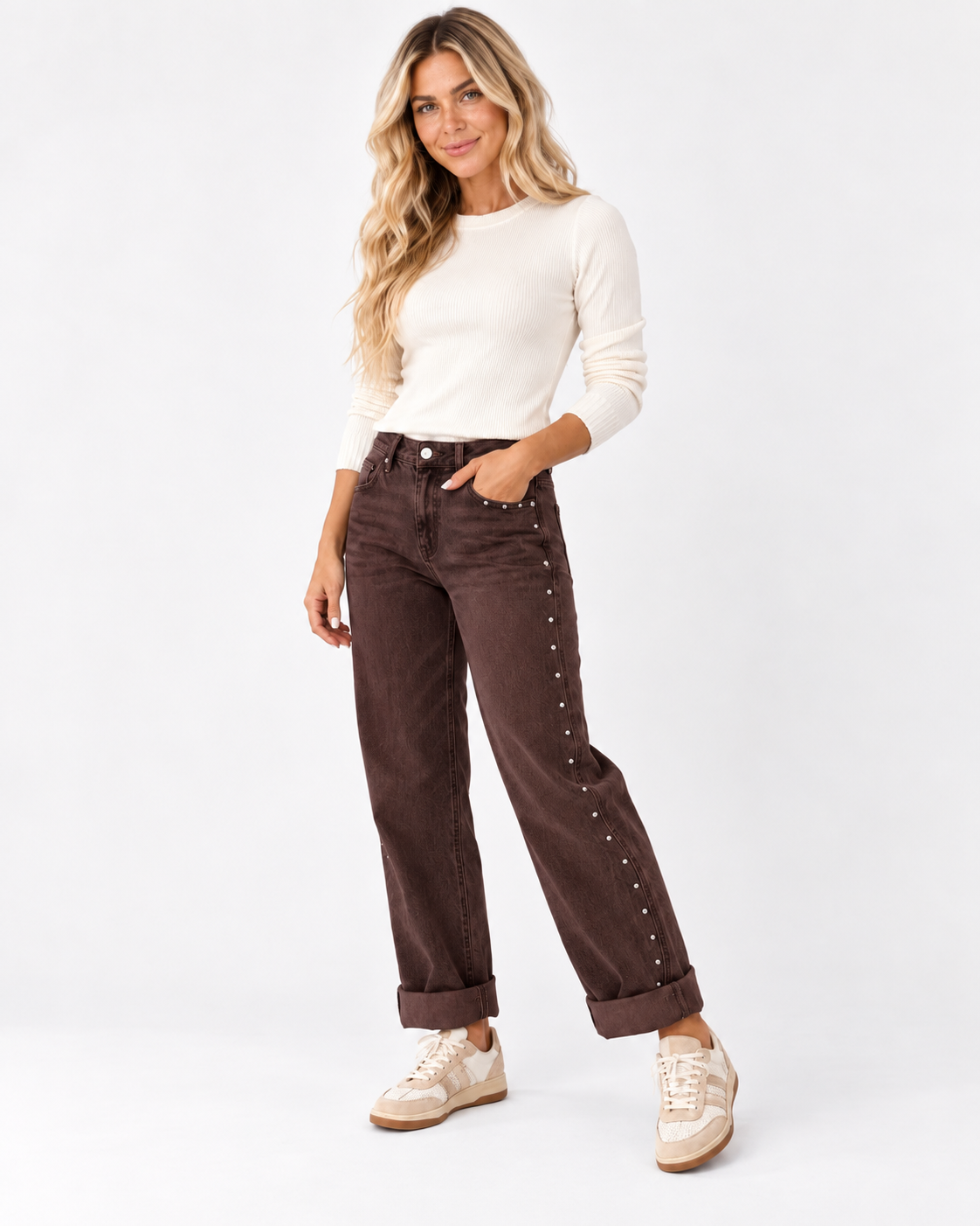 A woman modeling dark brown corduroy pants with silver stud embellishments down the side, styled with a cream long-sleeve top and beige sneakers against a white studio background.