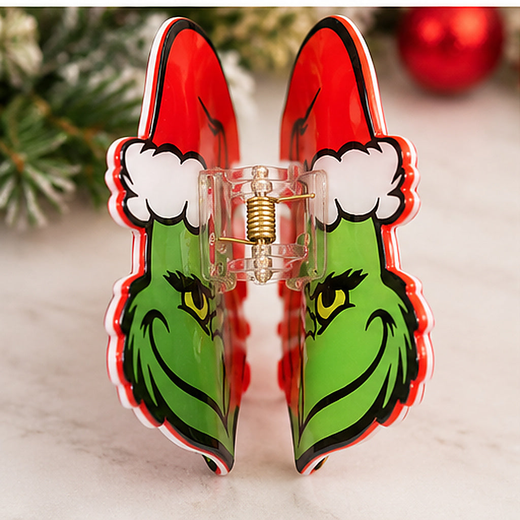 A novelty hair claw clip designed to look like the Grinch wearing a Santa hat, standing upright on a light marble surface with blurred Christmas greenery and a red ornament in the background.