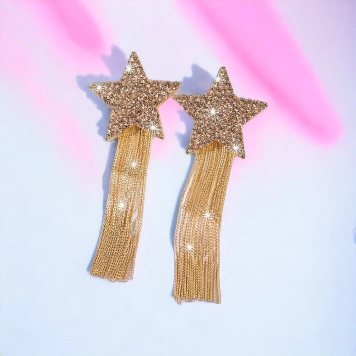 Gold star-shaped earrings with tassels on a pastel pink and blue background