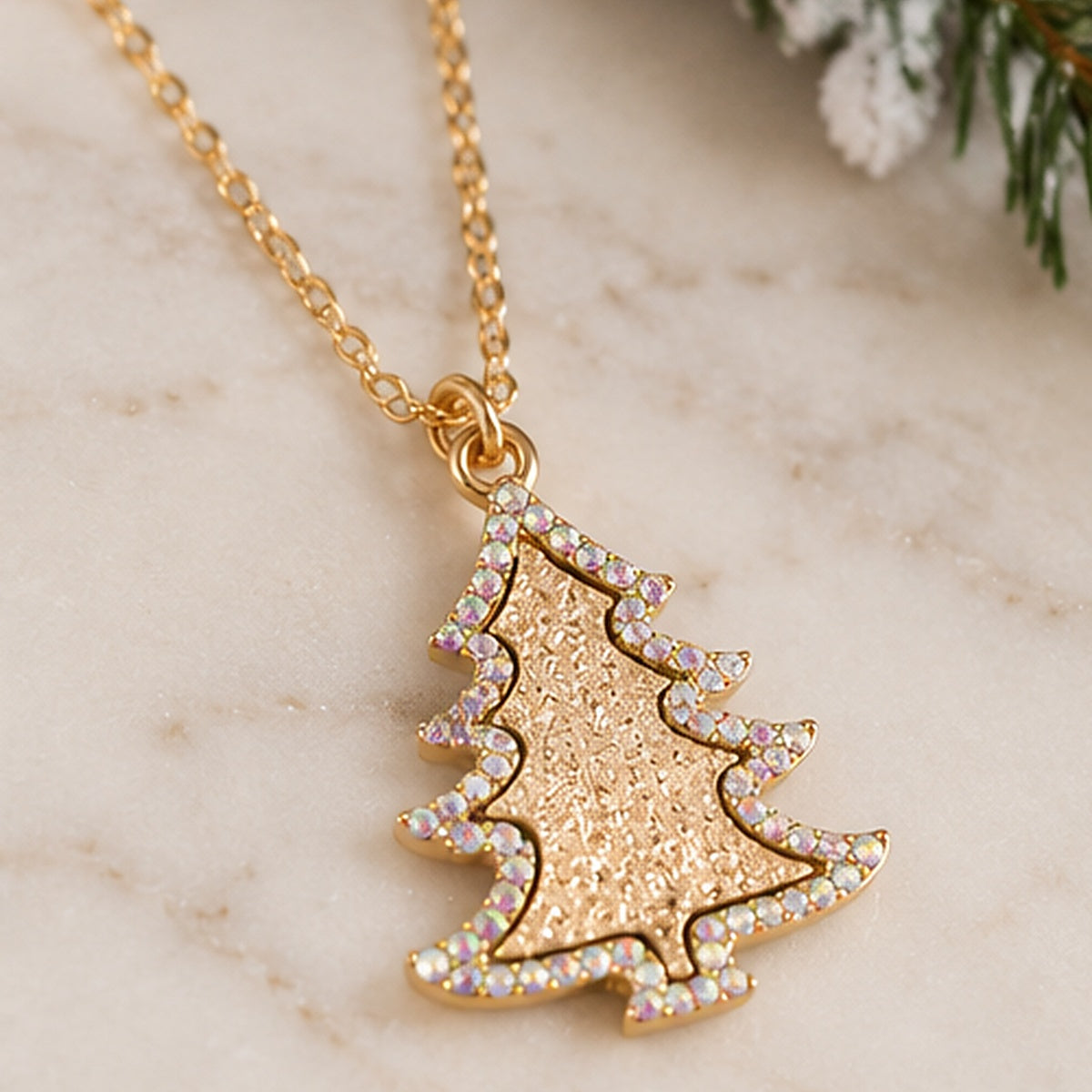 A gold Christmas tree pendant necklace adorned with iridescent rhinestones, resting on a light marble surface.