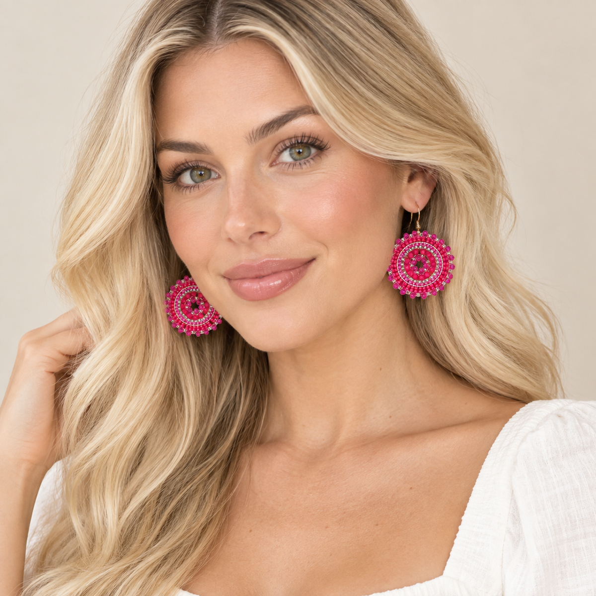 A blonde woman wearing large circular pink and red beaded drop earrings, photographed against a soft beige studio background.