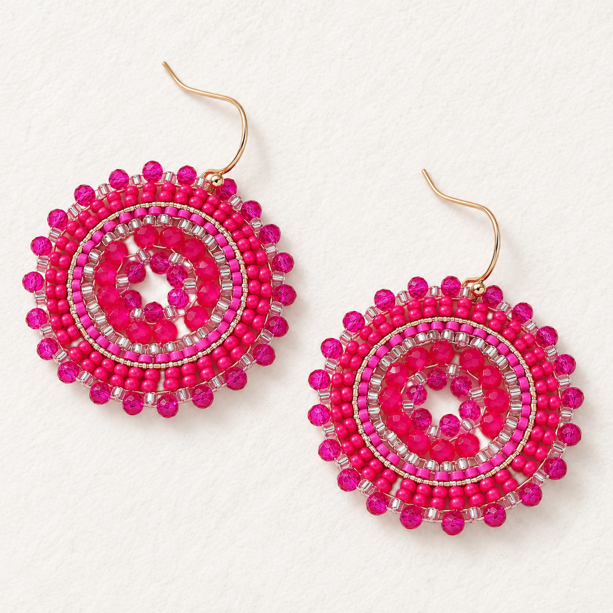 A pair of handmade circular dangle earrings featuring bright pink seed beads, clear rhinestone accents, and gold-tone fishhook wires, displayed on a textured off-white background.