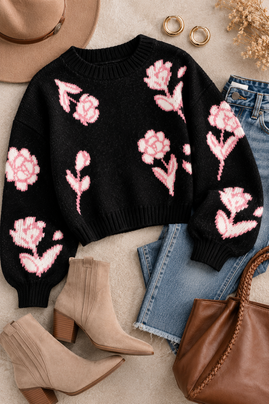A black knit sweater with pink floral patterns, styled in a flat lay with blue jeans, beige ankle boots, a brown handbag, a tan hat, and gold rings on a neutral textured background.