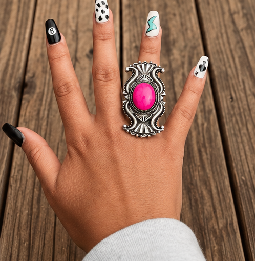 A hand wearing a large silver-toned ring with a bright pink oval stone, shown against a rustic wooden plank background. The hand has decorated nails in black, white, and mint green.