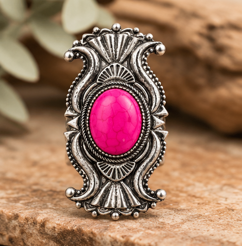An ornate silver-toned ring featuring a large, vibrant pink cabochon stone in the center, displayed on a textured wooden surface with a blurred natural background.