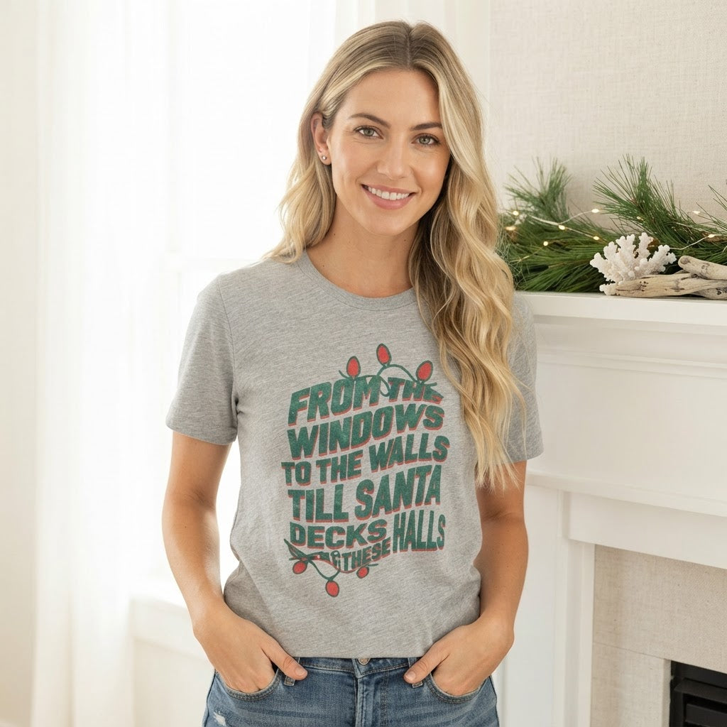 A woman wearing a heather gray graphic t-shirt with a Christmas-themed slogan printed in green and red, standing in a brightly lit indoor setting with a fireplace mantel in the background.