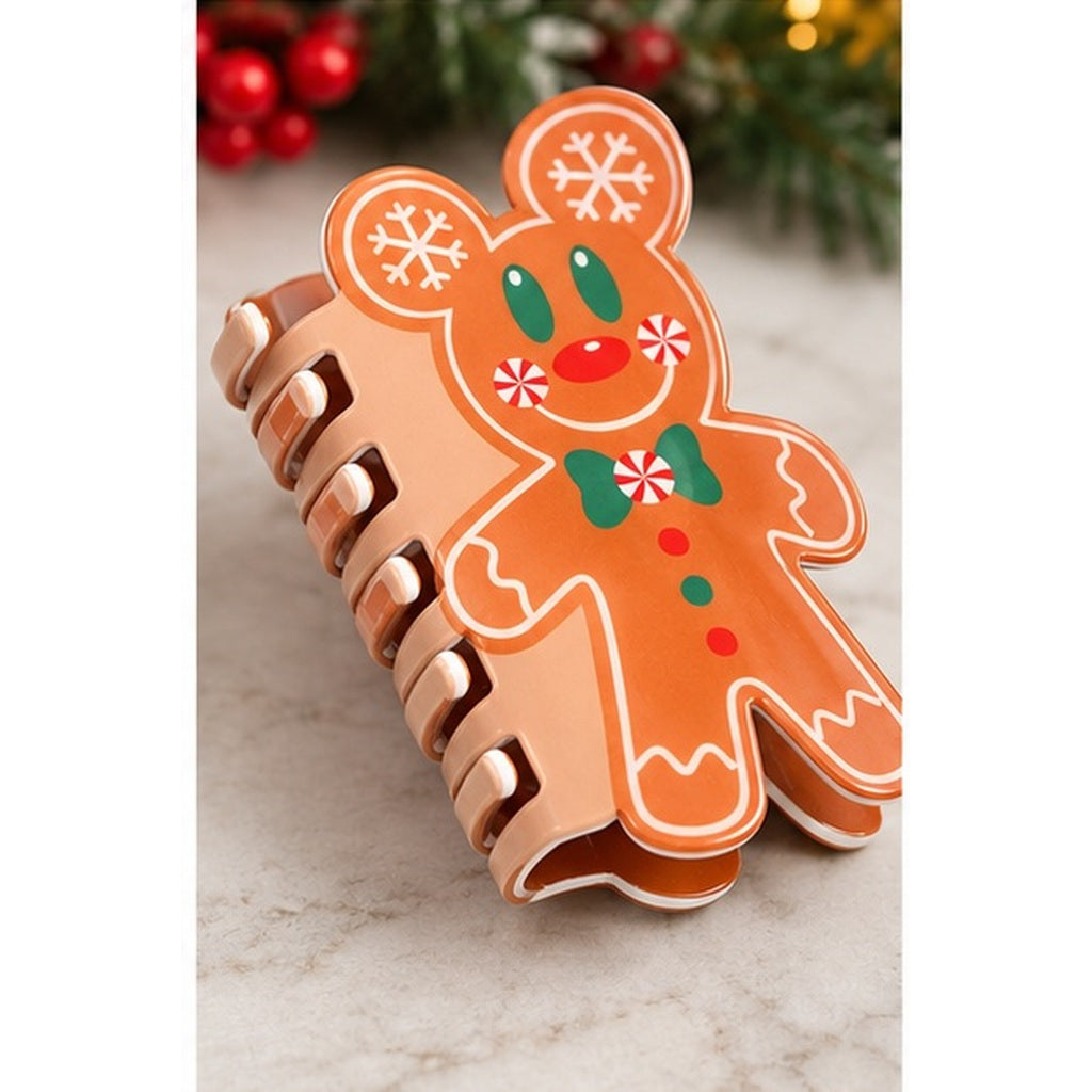 A spiral hair claw clip shaped like a gingerbread person, featuring an orange-brown body with white icing-style outlines, green bow tie and eyes, red nose and buttons, and white snowflake details on the ears, shown on a light marble surface with blurred holiday greenery and red berries in the background.