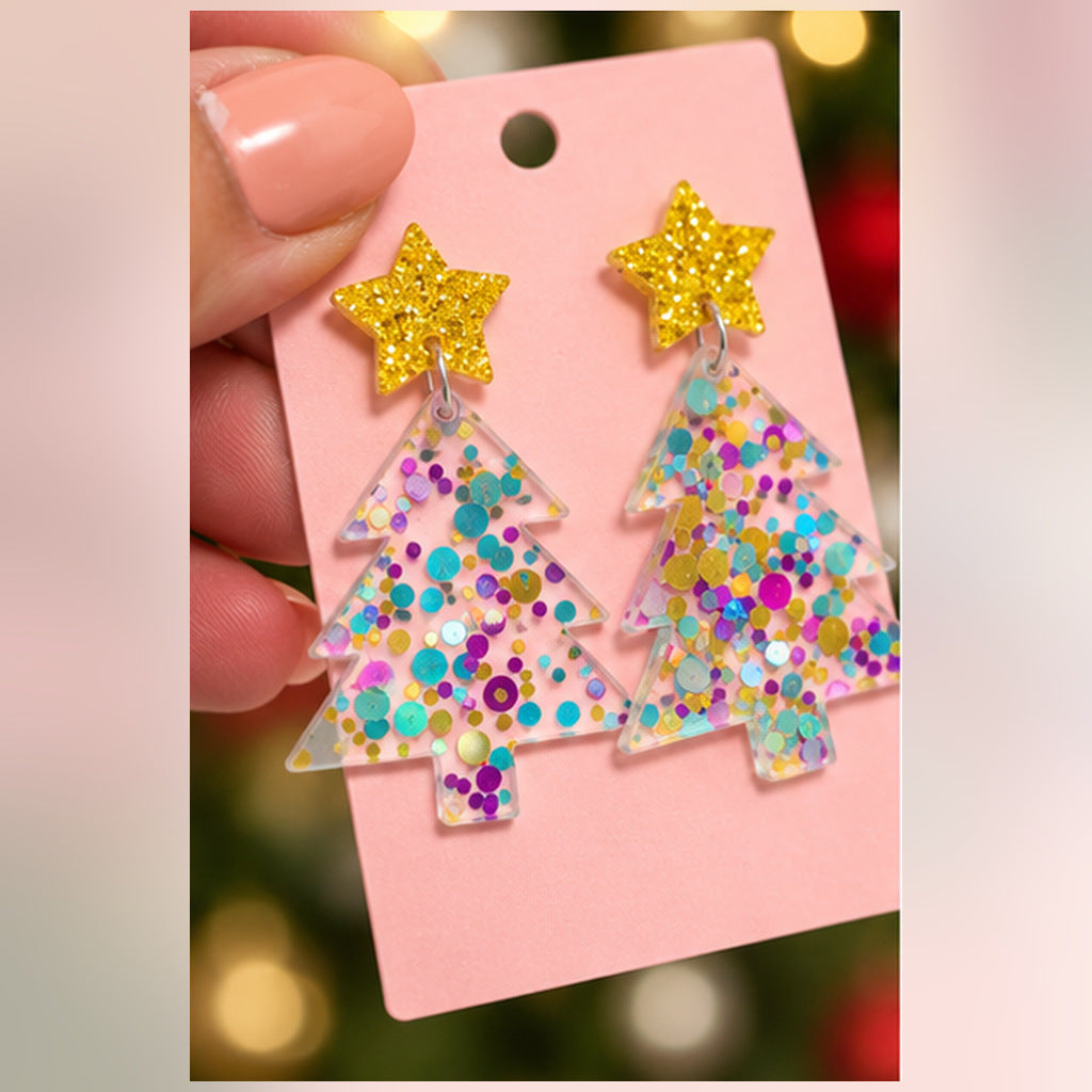 A pair of dangling earrings shaped like Christmas trees, made from clear resin filled with multicolored glitter, each topped with a glittery gold star. They are displayed on a pink backing card held by a hand against a blurred festive background.