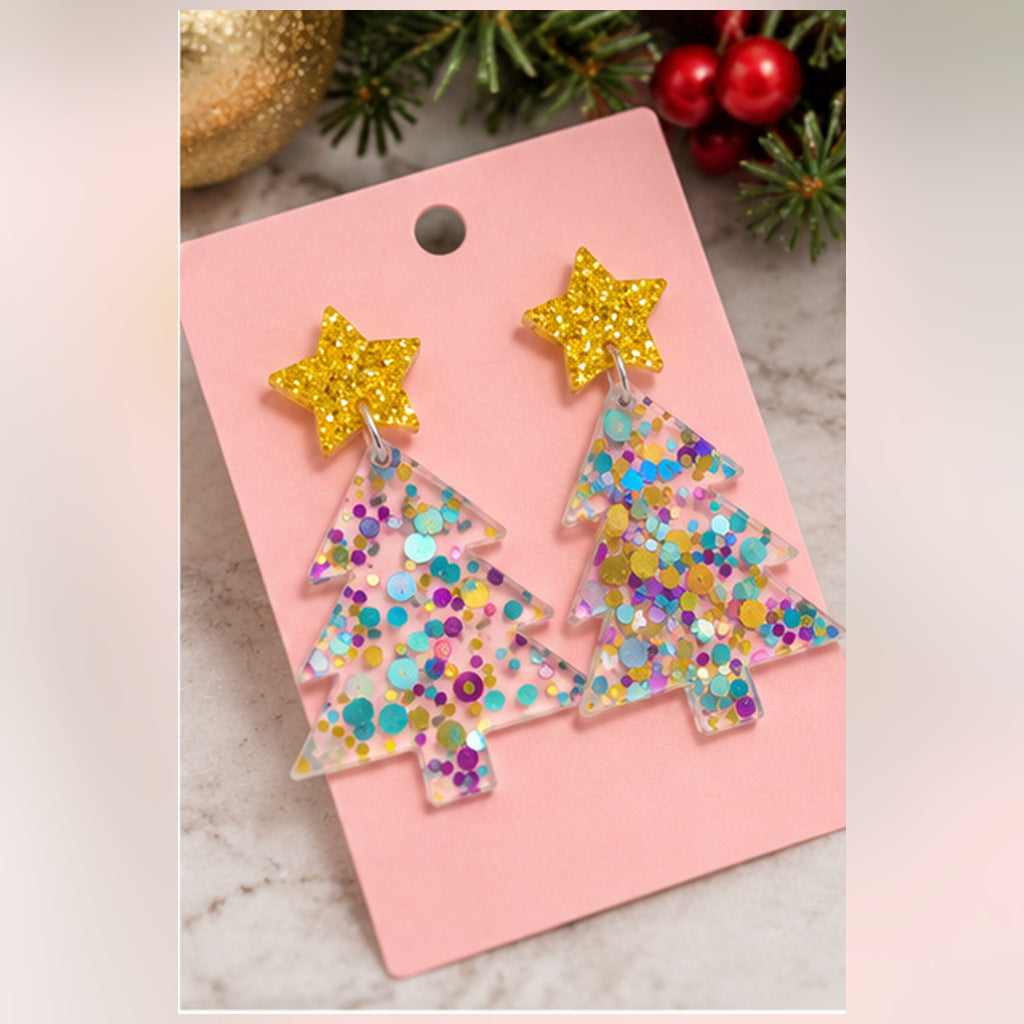 A pair of Christmas tree-shaped dangling earrings with glittery gold star tops and multicolor glitter tree bodies, displayed on a pink card against a holiday-themed background.