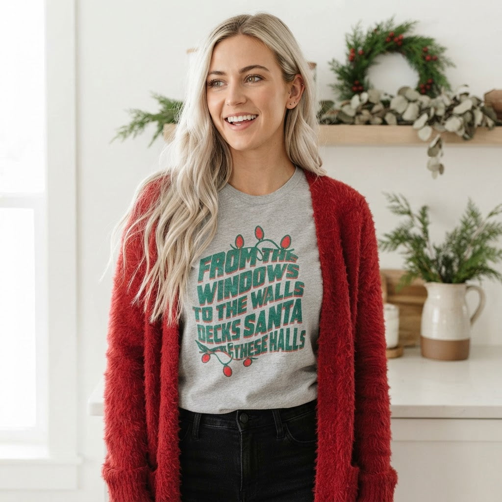 A woman wearing a gray graphic t-shirt with a Christmas message and a fuzzy red cardigan, standing in a bright, decorated indoor setting.