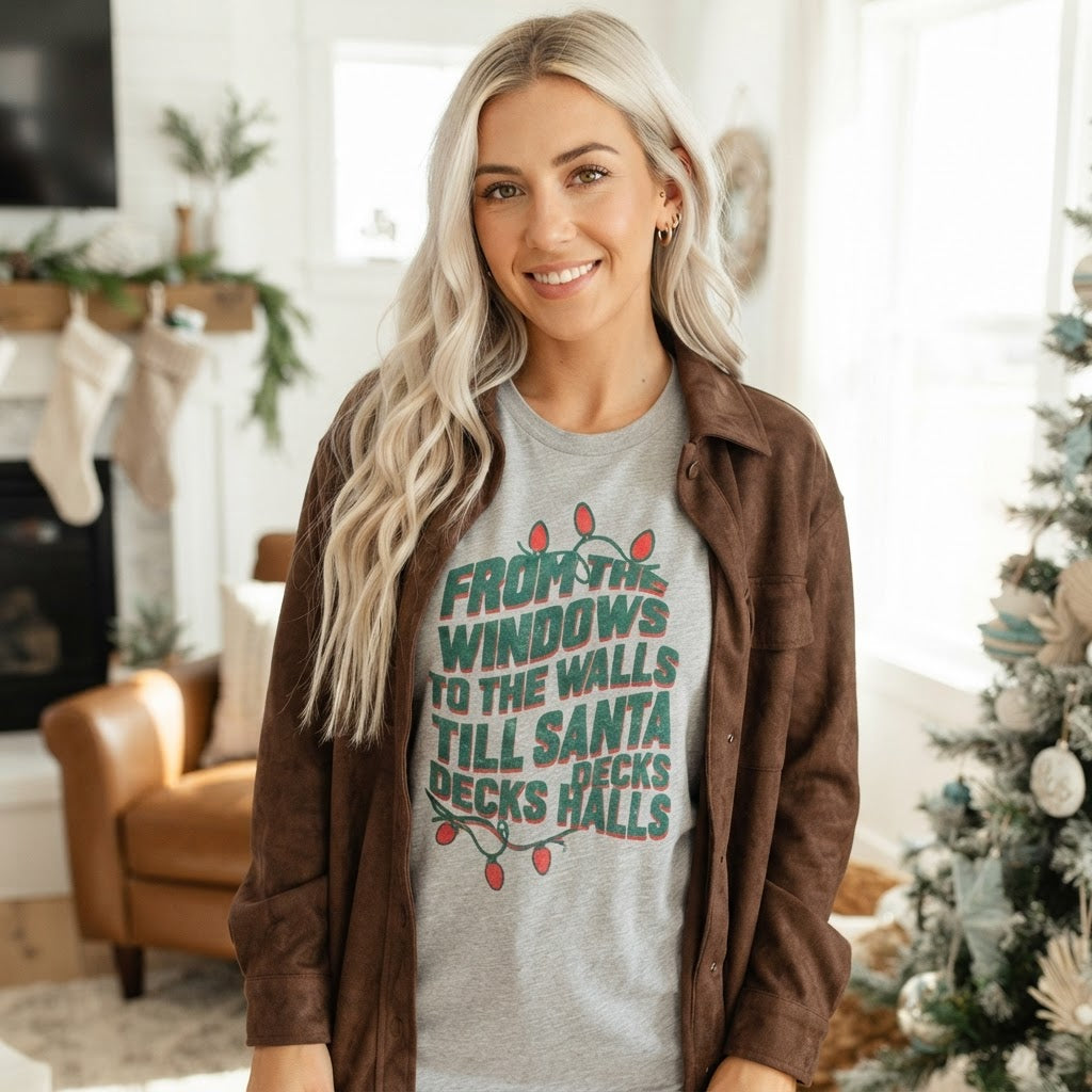 A woman with long blonde hair stands indoors wearing a heather gray graphic T-shirt with a Christmas-themed quote in green and red, layered under a brown button-up shirt. The background shows a softly blurred living room with a fireplace, chair, and decorated Christmas tree.