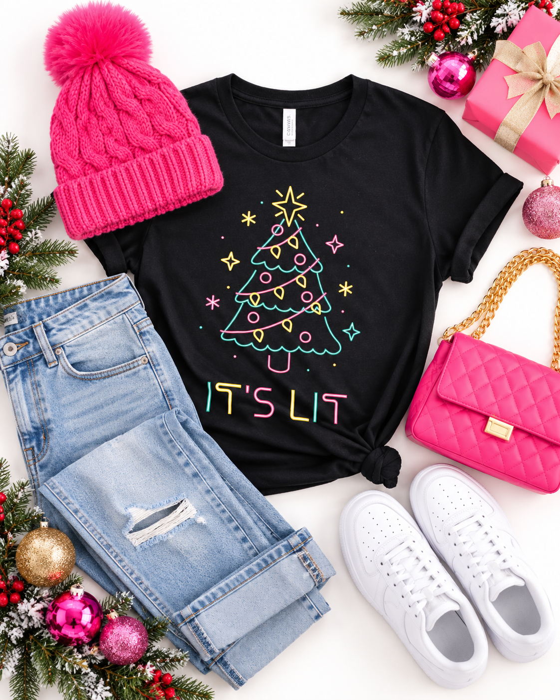 A styled holiday flat-lay featuring a black graphic t-shirt with a colorful Christmas tree and 'IT'S LIT' text, paired with a hot pink knit beanie, light-wash distressed jeans, a pink quilted handbag, white sneakers, and festive Christmas decorations on a white background.