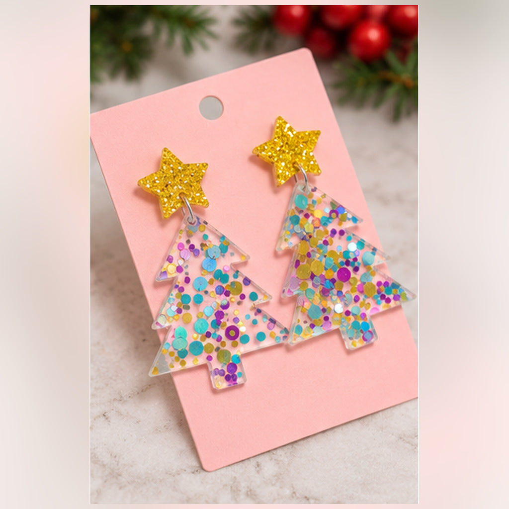 A pair of tree-shaped dangling earrings with glittery multicolor confetti and yellow glitter star tops, displayed on a pink card against a light marble surface with blurred holiday greenery and red berries in the background.
