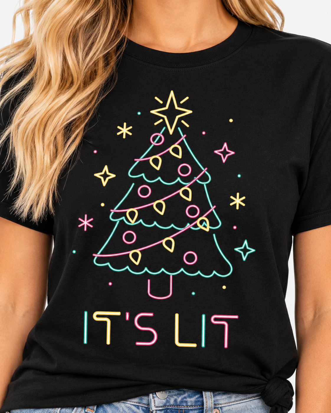 A woman wearing a black graphic t-shirt with a colorful neon Christmas tree design and the text 'IT'S LIT' in pastel pink, yellow, and blue. The image is cropped to show the torso, blonde hair, and the graphic print.