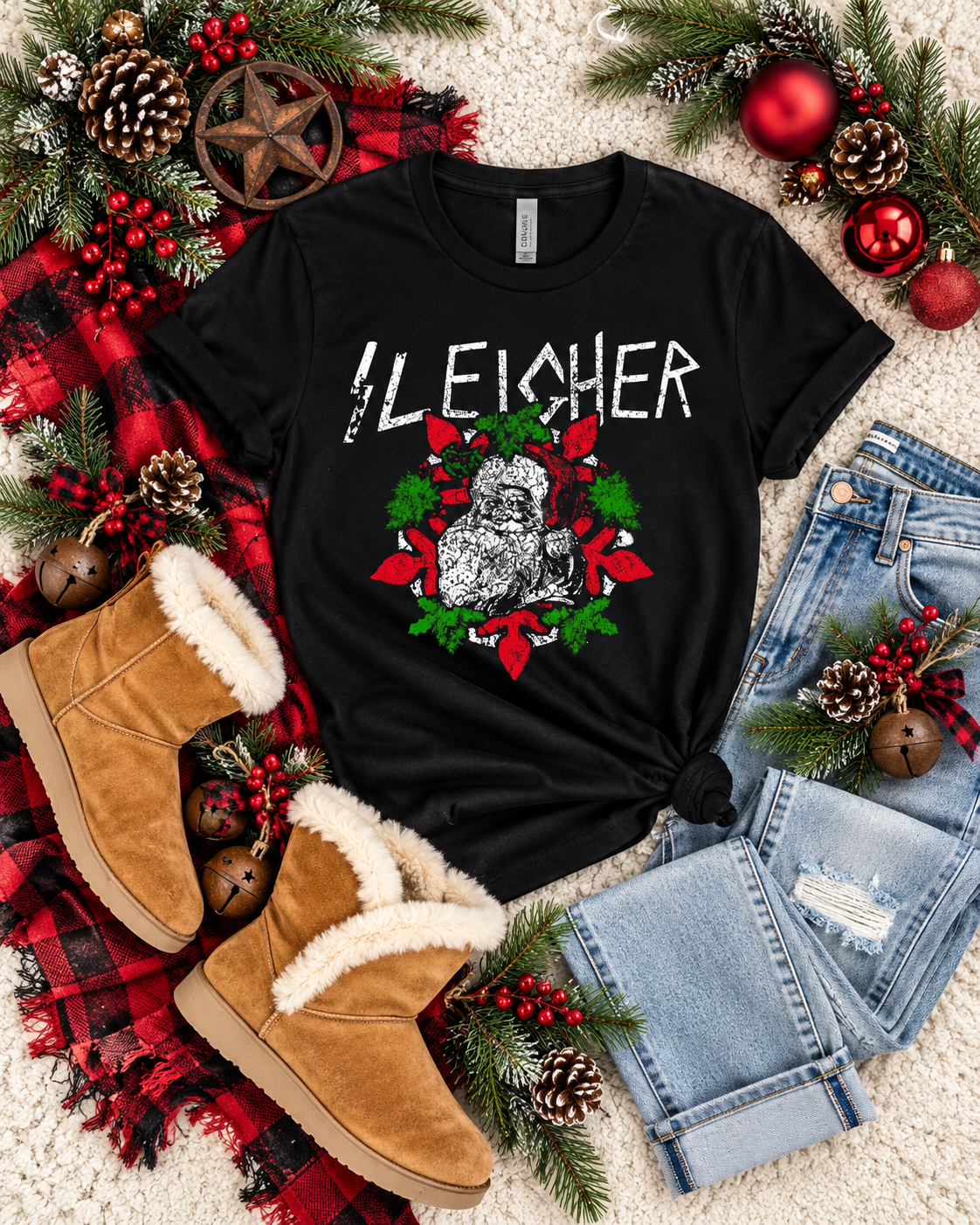 A black graphic t-shirt featuring a distressed white 'BLEACHER' text and a Christmas wreath design with a cat's face, styled in a flat lay with jeans, tan boots, and holiday decorations on a white carpet.