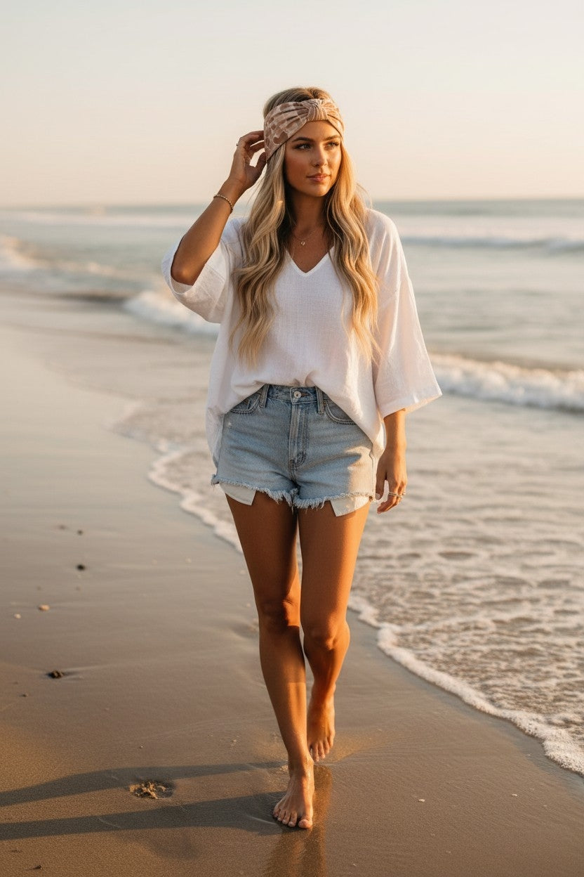 Woman walking on a beach wearing a white top and denim shorts.