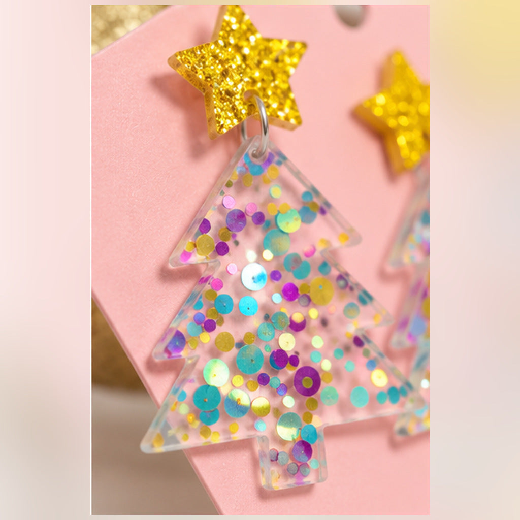 A close-up of a clear acrylic Christmas tree earring filled with iridescent confetti and sequins, attached to a glittery gold star stud, displayed on a pink card.