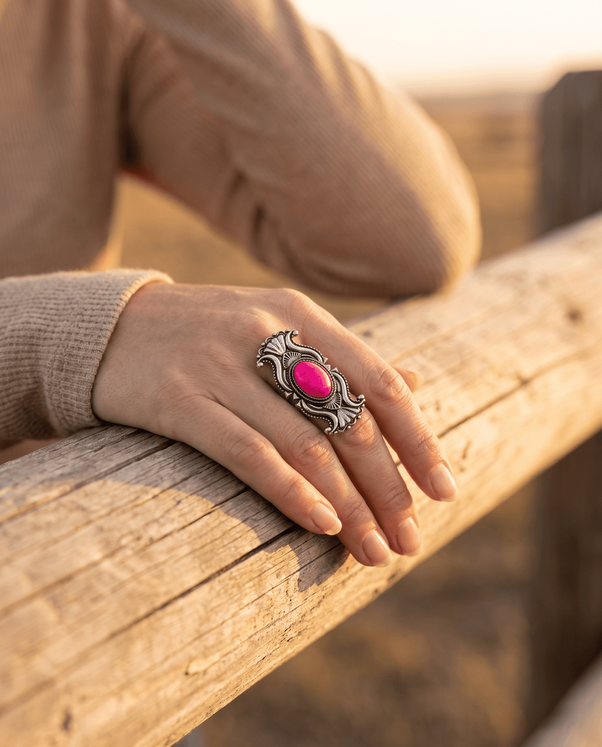 A close-up of a person's hand resting on a weathered wooden fence, wearing a large silver-toned ring with an oval bright pink cabochon stone.