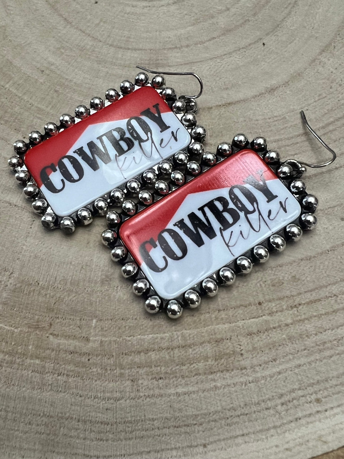 Pair of earrings with 'Cowboy Lifer' text on a wooden surface